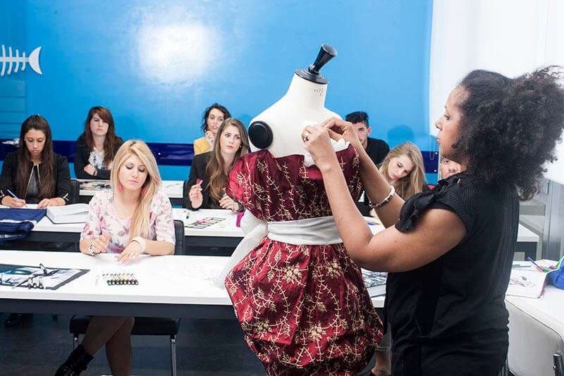 Docente al manichino con abito rosso ricamato davanti alla classe, aula design della moda AreaDomani