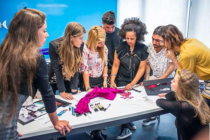 Gruppo di allievi con docente attorno a un tavolo con tessuti e figurini